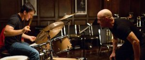 A scene from the movie 'Whiplash' featuring Miles Teller as Andrew Neiman intensely playing the drums, while J.K. Simmons as Terence Fletcher leans in, watching closely in a rehearsal room setting.