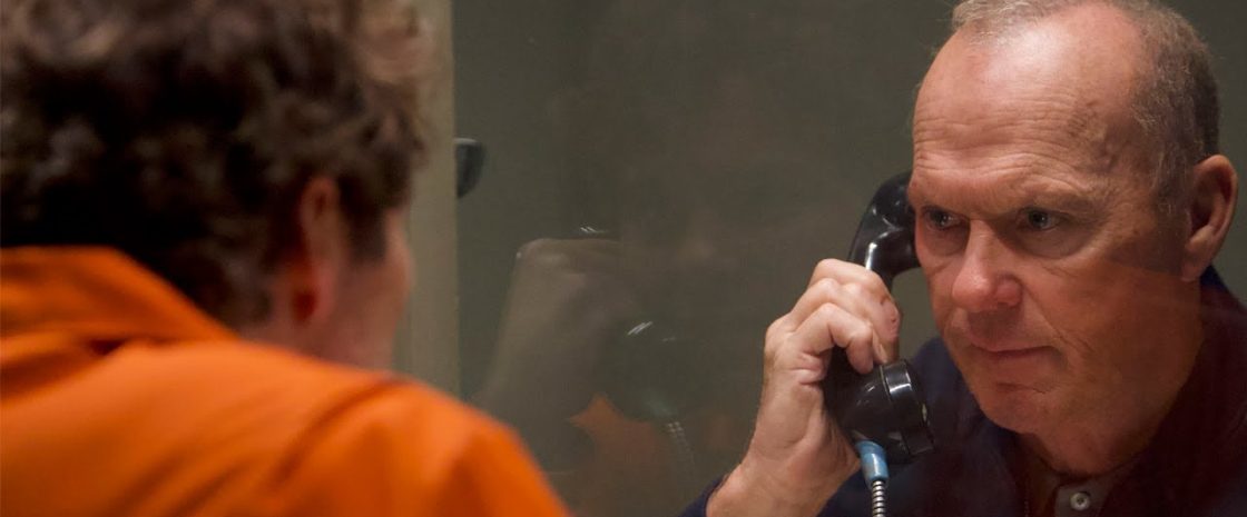 Michael Keaton in a tense prison visitation scene from Knox Goes Away, speaking through a phone to James Marsden in an orange jumpsuit.